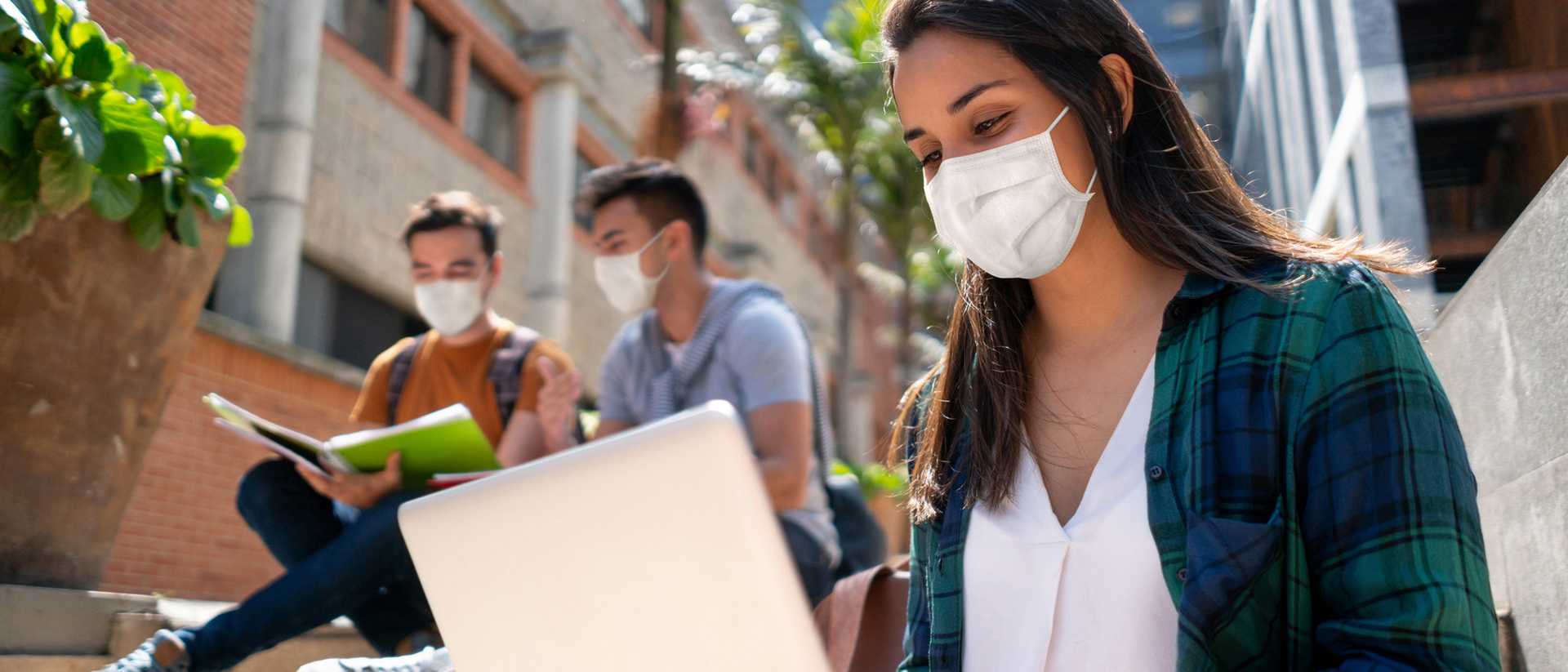 A woman with a face mask sits outside on a staircase with an open laptop on her lap. Two men sit behind her looking at a textbook.